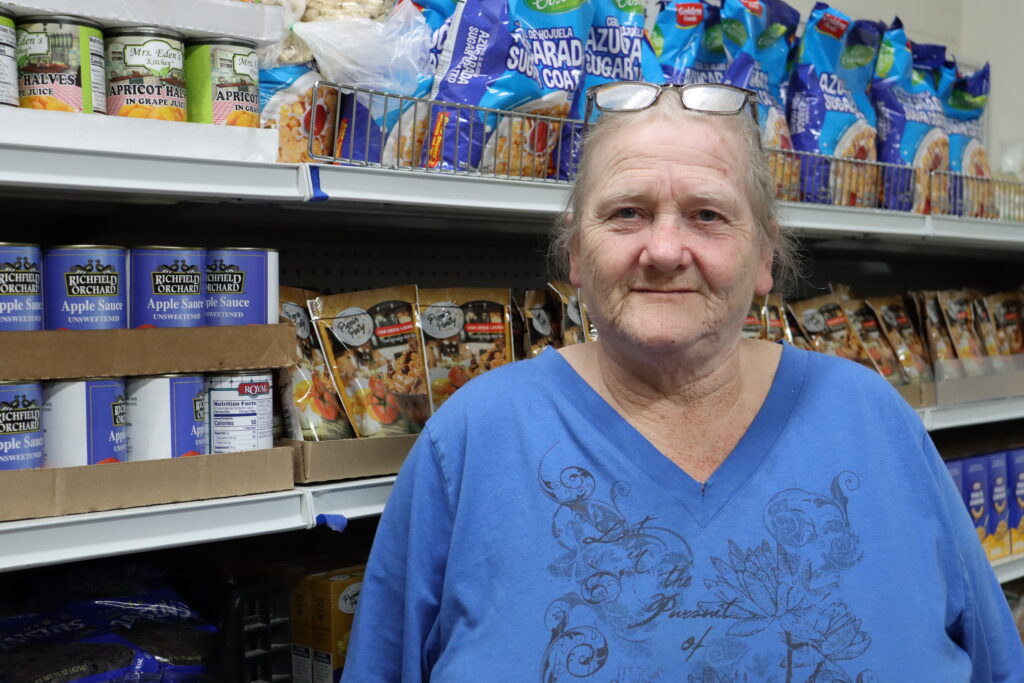 A woman (Jackie) stands in the TrueNorth Food Center in front of shelves stocked with food staples